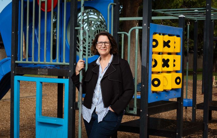 A woman wearing a dark jacket and striped shirt leaning against children's play equipment while smiling at the camera.