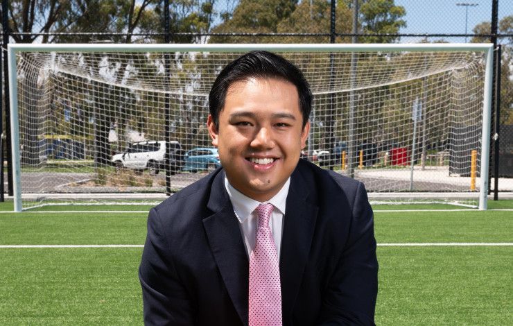 A man in a dark suit crouches on one knee on a synthetic soccer pitch while holding an orange soccer ball.