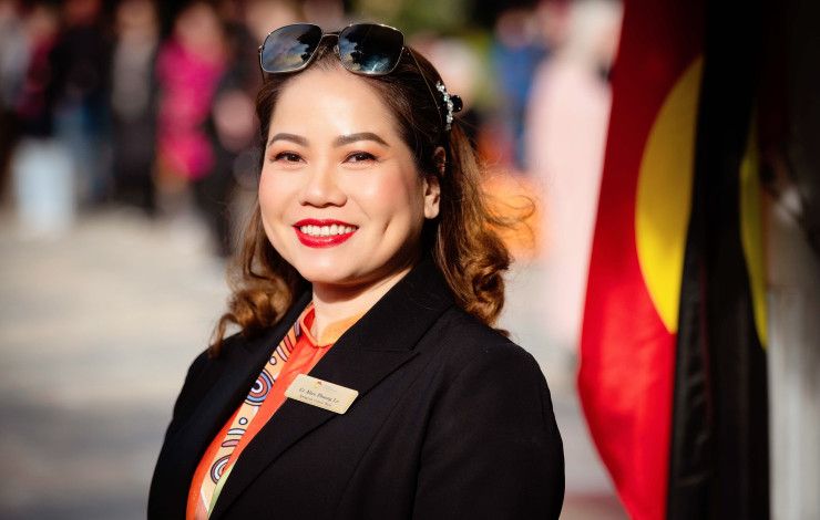 A smiling woman in a black jacket stands side on and smiles to the camera with an Aboriginal flag in the background.