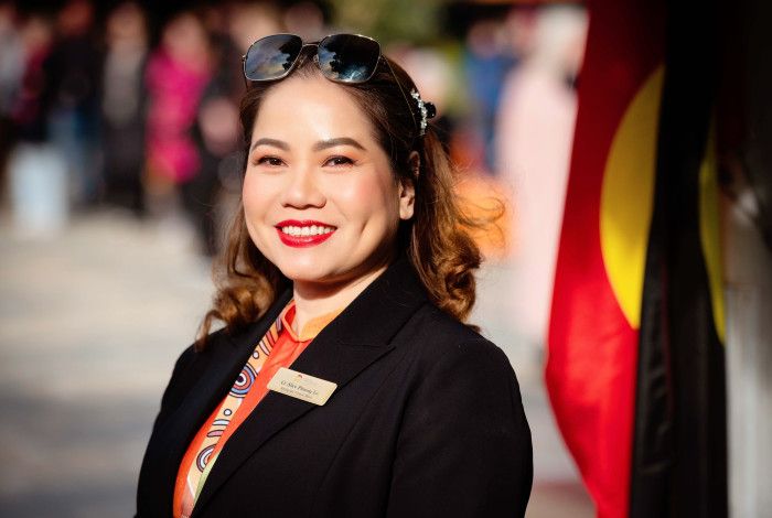 A smiling woman in a black jacket stands side on and smiles to the camera with an Aboriginal flag in the background.