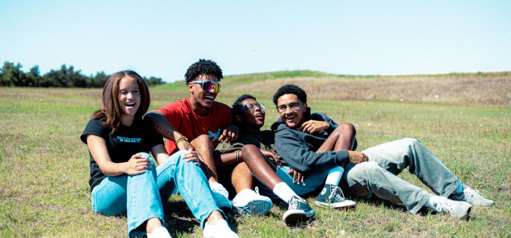 Four young people sitting together and laughing on a grassy hill.