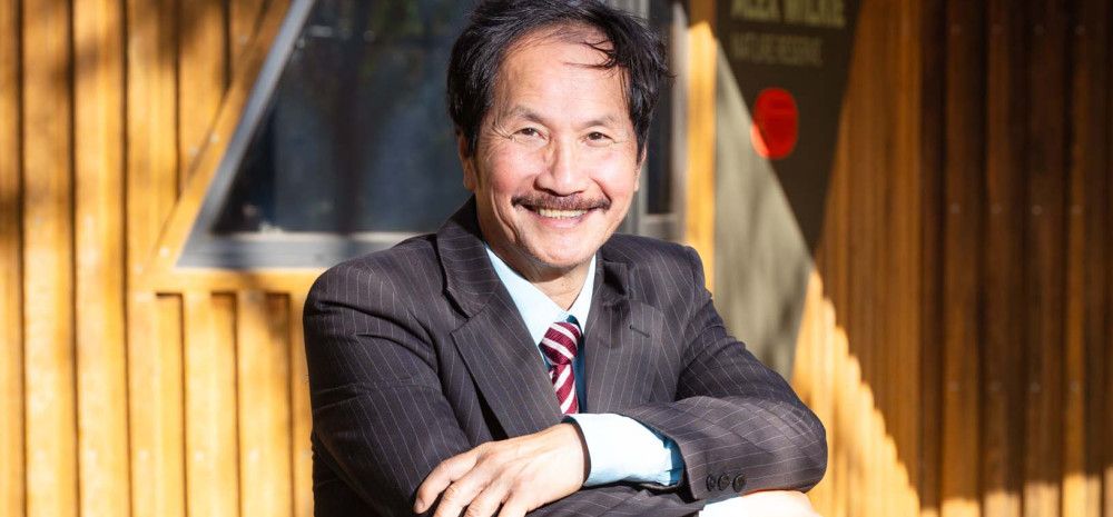 A man with dark hair and a moustache smiles at the camera with his arms crossed in front of him