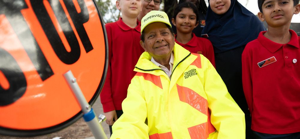 A smiling man dressed in high vis holding a stop sign while surrounded by smiling school children