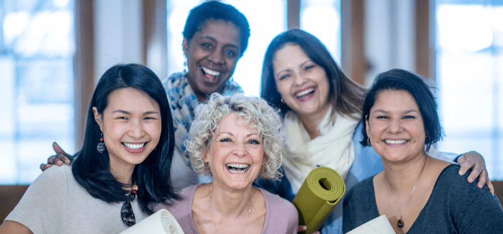Five people of different ages and cultural backgrounds are standing together in a group. They are smiling and holding yoga mats.