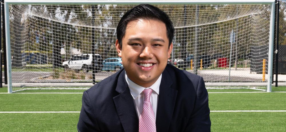 A man in a dark suit crouches on one knee on a synthetic soccer pitch while holding an orange soccer ball.