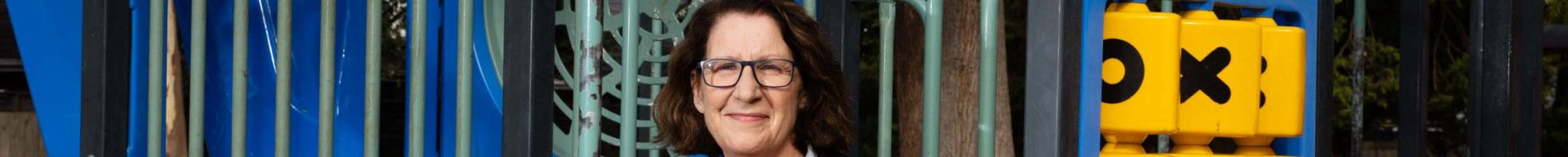 A woman wearing a dark jacket and striped shirt leaning against children's play equipment while smiling at the camera.