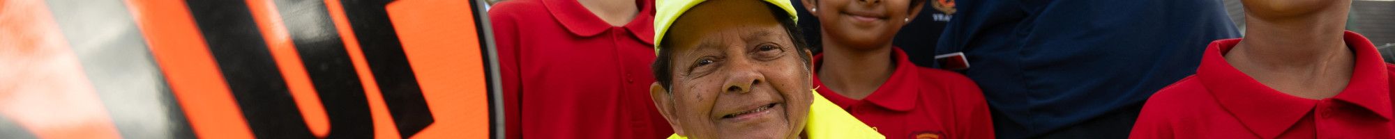 A smiling man dressed in high vis holding a stop sign while surrounded by smiling school children