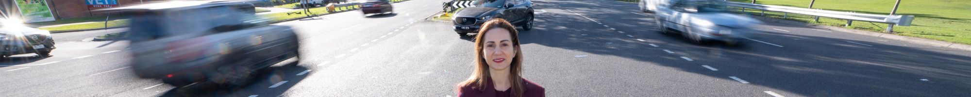 A woman dressed in a suit standing on a grass island in the middle of a busy road.